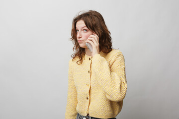 Young woman with curly hair wearing yellow knitted sweater talks on smartphone. Isolated on light...