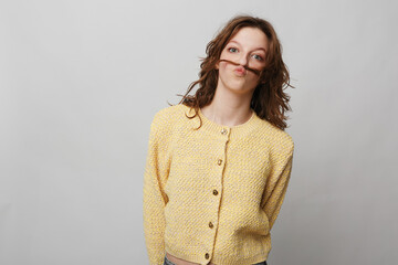 Obraz premium Young woman in yellow knitted sweater makes a playful mustache with her hair, standing against a plain light background. Concept of fun, humor, and casual lifestyle portrait.