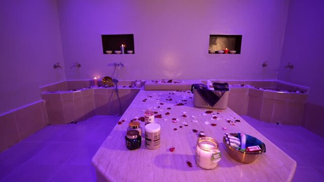Traditional Moroccan hammam spa interior with marble table, rose petals, candles, running water, and bath products under relaxing purple ambient lighting.