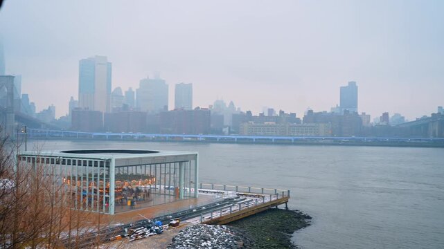New York, USA, 25 December 2025: Glass carousel building on snowy Brooklyn waterfront in fog. Glass enclosed carousel on a pier with snow and foggy Manhattan skyline in the background