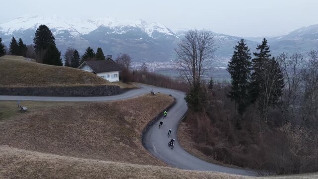 Static drone shot facing a narrow alpine road with a sharp hairpin turn during early morning blue hour. Four cyclists with bike lights descend the winding mountain road while a wide valley and snow-co