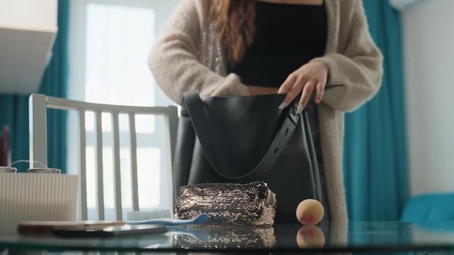Caucasian Woman Packing Foil Lunch And Peach On Glass Table, Placing Reusable Container And Napkin Into Black Tote, Sunlight Through Curtains, Quick Morning Routine, Casual Cardigan Commuter Ready