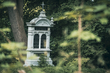 Zabytkowy, bogato zdobiony grobowiec w formie klasycystycznej kapliczki na starym cmentarzu. Widok przez zielone liście na monumentalny nagrobek w cieniu drzew. © Henryk Niestrój