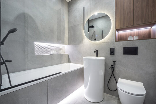 A bathroom with a bathtub and shower combination, a round mirror on the wall, and a black and white color scheme. There are modern fixtures and clean lines