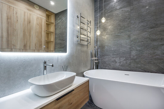 A bathroom features a freestanding tub and a modern sink on a wooden cabinet. The wall has gray tiles, and there is a mirror with lighting above the sink