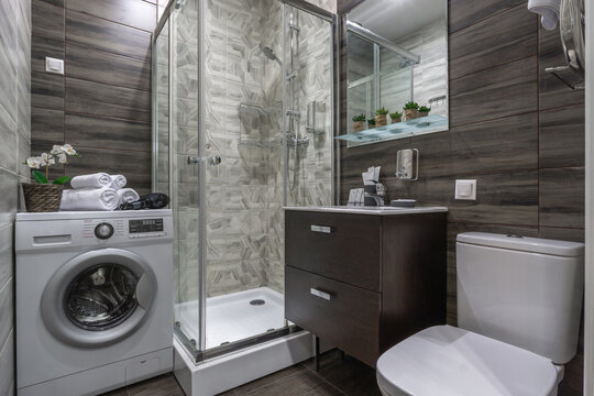 This small bathroom features a glass shower, a washing machine, a sink with storage, and a toilet. It has simple decor with towels and small plants on the shelves