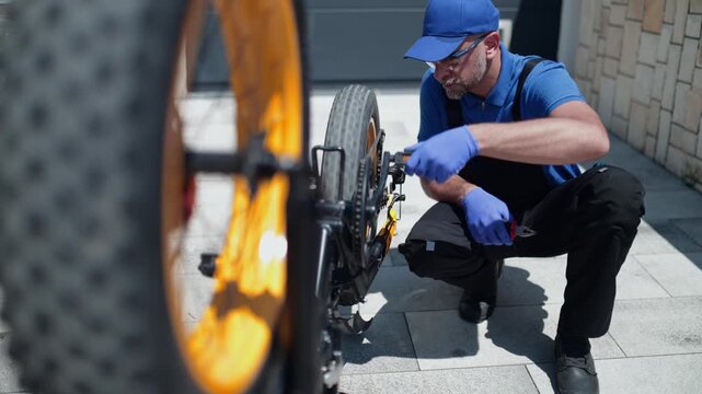 Close-up video of a bicycle mechanic in blue workwear and protective gloves repairing the pedal and drivetrain of an electric fat tire bike outdoors in bright daylight.