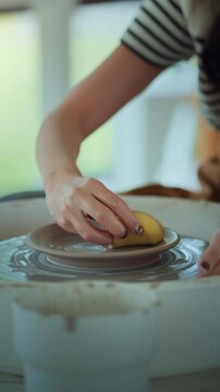 Close-up of hands of female artist shaping wet clay on spinning pottery wheel with sponge &ndash; creative ceramic making process in modern craft studio, traditional handmade sculpting technique