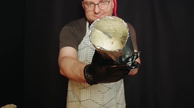 Caucasian man with glasses wearing apron and red bandana holds foil lined tin. His gloved hands presenting batter container against black background. Preparing ingredients for baking bread in a pan
