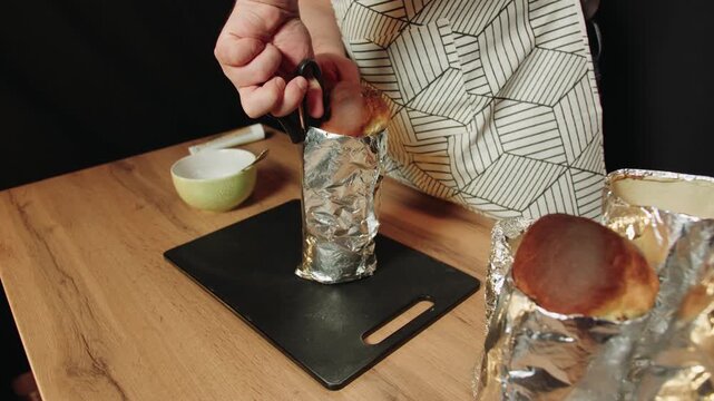 Pastry chef uses kitchen scissors to cut through a foil pan to reveal a freshly baked loaf of bread on a cutting board on a wooden table. A man in an apron prepares bread for eating.
