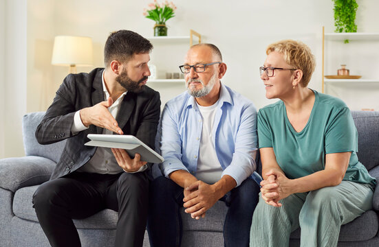 A real estate agent reviews contract details on tablet while mature couple listens on their couch during a home consultation. Male advisor consulting seniors about investments and insurance. 