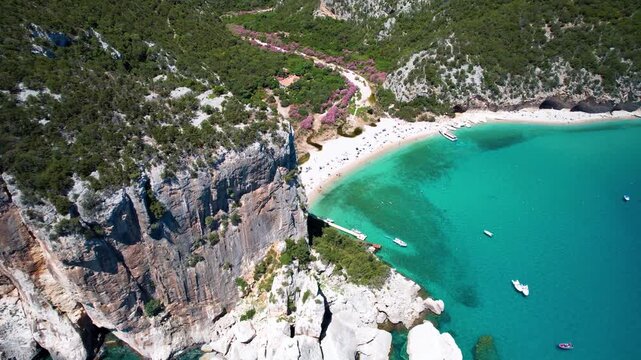 Sardinia Cala Luna beach aerial view reveal behind high cliff with tourist boats on turquoise sea Golfo di Orosei Italy white sand coast drone footage 4k cinematic landscape travel destination