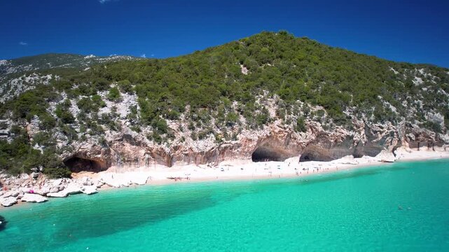 Cala Luna Beach Side Slide Aerial Drone Video. Three Huge Caves on White Sand with Tourist Boat. Sardinia Italy. Turquoise Sea and Limestone Cliffs European Caribbean