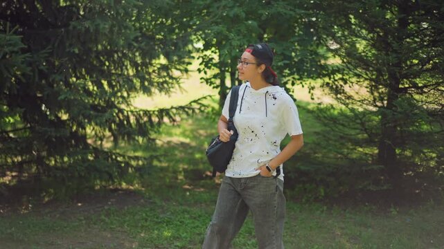 Latina explorer pausing beneath green canopy, handsonhips survey of parkland, confident stance with shoulder bag, dappled light through branches, casual summer outfit, observational mood and candid