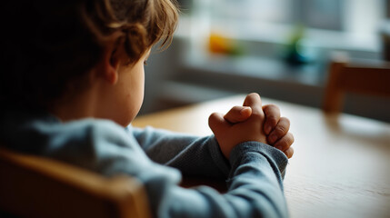 Faceless child from behind at a simple table or chair in a plain interior setting small hands clasped together on the surface in a prayer posture soft clean light from a nearby
