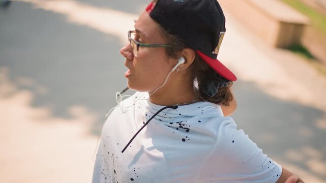 young person in cap walking, listening to music with earbuds, wearing glasses and backward cap, relaxed head movements, sunlit sidewalk, summer breeze, casual tshirt, expressive profile, leisurely