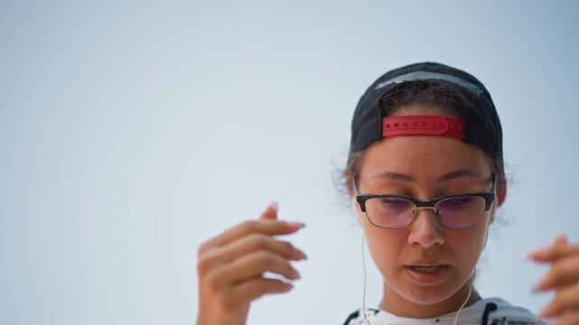 closeup young adult adjusting earbuds sky, checking smartwatch and cord, fitting earphones with concentrated expression, cap worn backward, glasses, preparing for outdoor run, soft morning light,