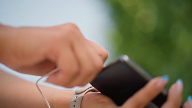Commute closeup caucasian woman hands with smartphone connecting wired headphones in shaded park, white cable threading over fitness wristband, focused fingers with neat manicure, soft bokeh, short
