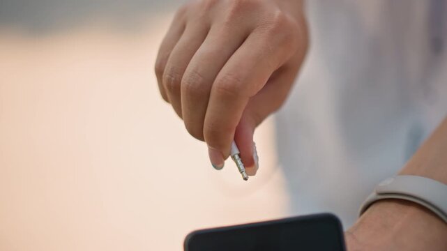 closeup hands plugging earbud into smartphone, female jogger wearing white smartwatch, warm sunset bokeh, careful finger motion inserting .mm jack into port, cable looped over phone, soft fitness