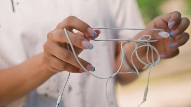 closeup hands untangling knotted white earphones, student musician works through cable snarl with patient fingers, sunlight highlights nails and texture, park background, moment of relief as cord
