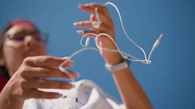 hands untangling white earbuds against blue, closeup outdoor shot showing wristwatch, casual shirt, focused fingers working through knot, commuter preparing for transit, sunlit warm tones, texture