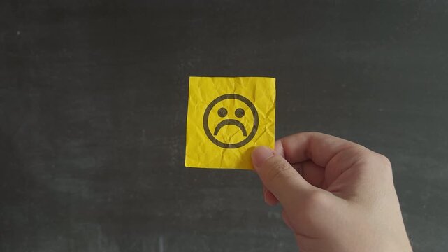 A person holding a wrinkled yellow note with a sad emoticon on it, symbolizing negative emotions. Close up.