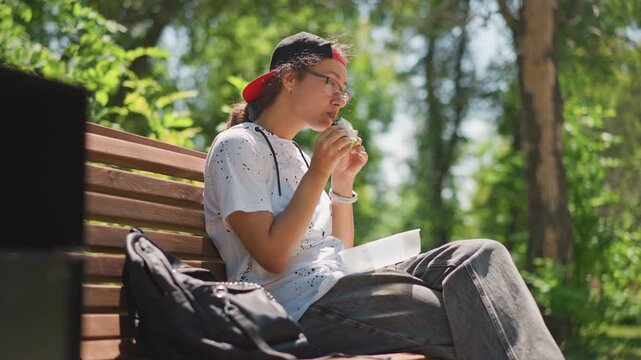 young woman nibbling snack on bench, side profile composition with sunlight, focused on notebook and page, backpack at side, tree canopy casting dappled shadows, concentrated study mood