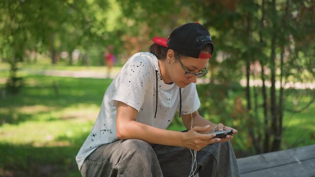 asian teen gamer leaning over phone focused in park, fast thumb movements and concentrated posture, earbuds in, cap backwards and backpack at side, dappled sunlight and leafy backdrop emphasize