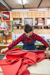 Naklejka premium Woman artisan measuring red fabric with a tape measure on a wooden table, preparing it for a custom upholstery project. Celebrating craftsmanship and small business