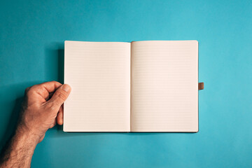 Obraz premium Top view of hand lifting notebook sheet against teal backdrop, simple stationery scene with soft light and modern minimal style.