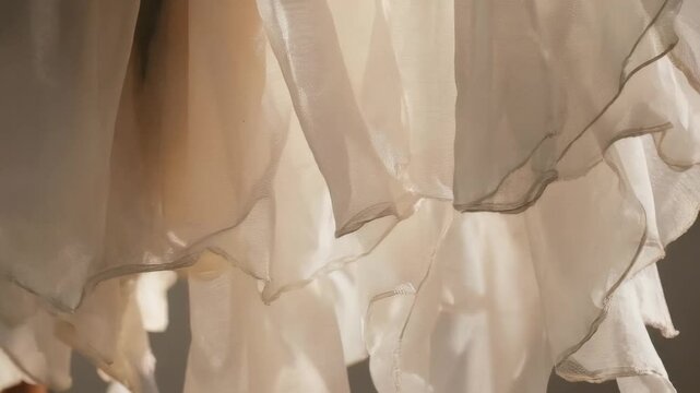 Sunlit chiffon layers cascade in soft, translucent waves, their scalloped hems glowing with warm highlights while gentle shadows reveal airy texture and delicate movement in a close-up frame.