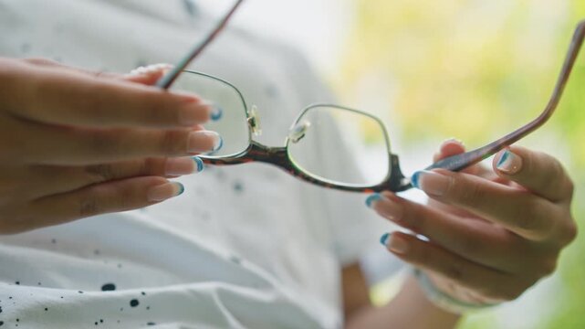closeup hands holding eyeglasses outdoors sunlight calm gentle scene of person adjusting tortoiseshell frames with turquoise manicure, white speckled shirt, soft bokeh background, tactile focus