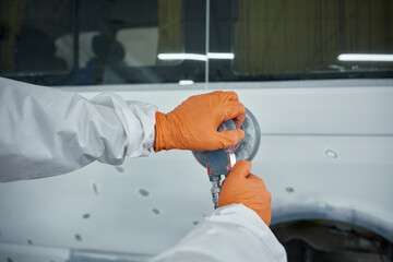 An auto body technician sands a car panel in a protective suit and orange gloves