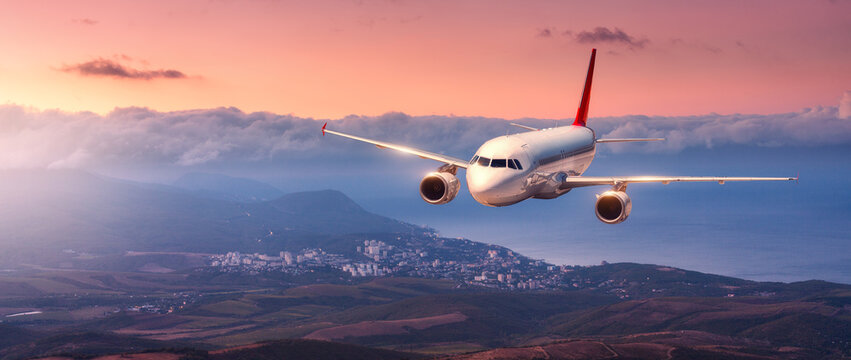 Commercial passenger airplane flying over sea coast and mountains at sunset. White aircraft in flight above sea and clouds during golden hour. Aviation, travel, transportation concept. Aerial view