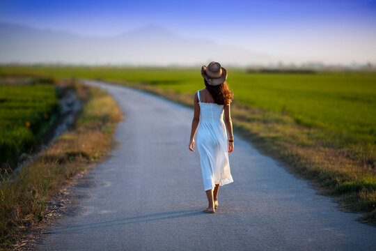 Young woman with a hat and white dress walking along a lonely path at sunset