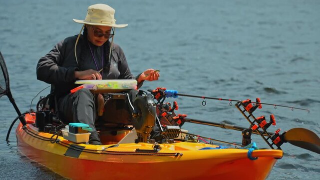 Kayaking lake sport fishing. Tourist enjoys kayak fishing at scenic coastline. Peaceful water setting at lake. Woman prepares her tackles lure or bait to the braid line while slowly floating on water