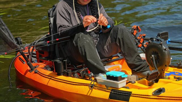 Kayaking lake sport fishing. Tourist enjoys kayak fishing at scenic coastline. Peaceful water setting at lake. Woman prepares her tackles lure or bait to the braid line while slowly floating on water