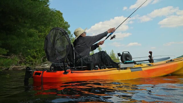 Woman kayaker moving on water waves and kayak fishing, casting and reeling under summer afternoon sun. Surrounded by green shoreline and the quiet nature, perfect balance concept of sport and solitude