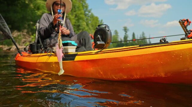 Kayaking lake, woman showing her casting pike lure while floating on kayak boat. Kayaking lake with scenic landscape. Woman angler in a sun hat on motorized kayak enjoying peaceful summer vacation.