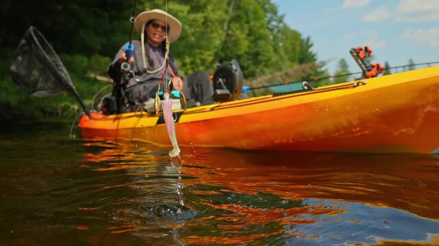Kayaking lake, woman showing her casting pike lure while floating on kayak boat. Kayaking lake with scenic landscape. Woman angler in a sun hat on motorized kayak enjoying peaceful summer vacation.