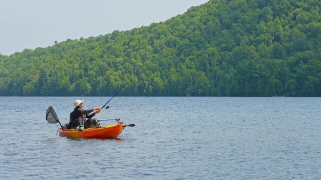 Woman casting and reeling while floating on kayak boat. Kayaking lake with scenic landscape.  Woman angler in a sun hat on motorized kayak enjoying peaceful summer vacation sunny day, active outdoor.