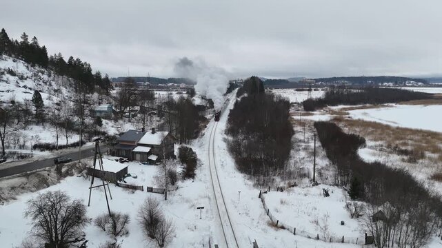 A steam train moves along tracks surrounded by snow and mountains. The train passes houses and fields under a cloudy sky during winter. Smoke rises from the engine as it travels"