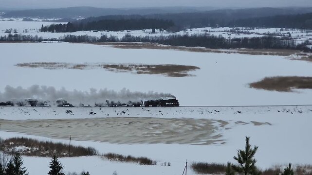 A steam train moves along a snowy area next to a frozen lake. The landscape shows white snow and tall trees in the background. The scene captures winter life in the countryside