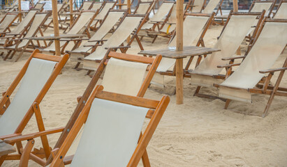 Rows of empty wooden beach lounge chairs with fabric seats arranged on sandy beach terrain under sunny conditions, ideal for summer vacation and relaxation themes © John_Doo78