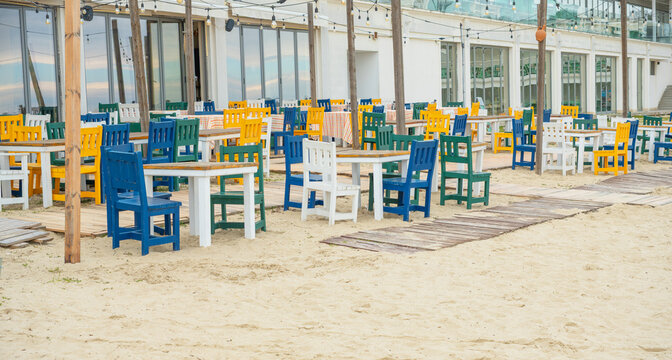 Colorful wooden chairs and tables arranged on a sandy beach terrace of a seaside restaurant, creating a vibrant and inviting atmosphere for summer dining