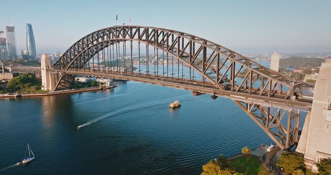 Sydney Harbour Bridge creating an iconic urban landscape with a ferry and sailboat navigating the deep blue water bay, framed by historical towers and modern city architecture Aerial drone panorama