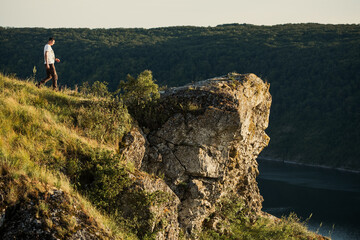 Young man hiking on rocky cliff edge overlooking river and forest landscape during golden hour sunset ideal for travel and adventure themes. © ninelutsk