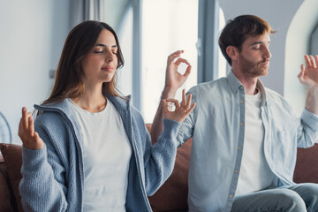 Fototapeta premium A calm couple sits on a sofa at home practicing meditation together, breathing slowly in a peaceful moment while focusing on mindfulness, emotional balance, and mental wellbeing