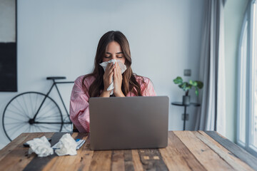 A frustrated young woman sits at home feeling ill with cold and flu symptoms, holding a tissue to...