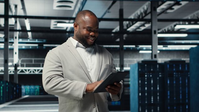 Smiling admin using tablet in in data center maintaining infrastructure for workloads. Joyful IT professional in server farm utilizing high tech server clusters for parallel processing, camera B